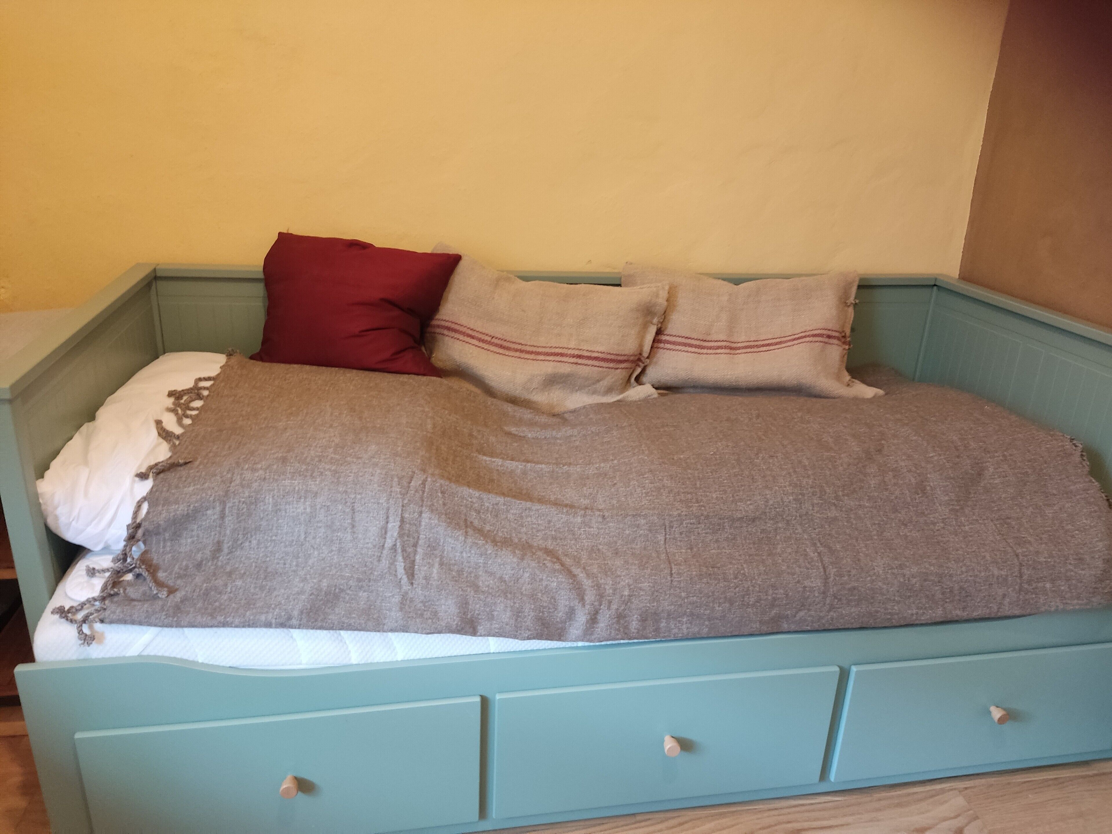 Ein Bett mit einer grauen Decke, zwei Kissen und einem roten Kissen.