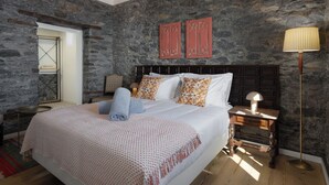 Une chambre avec un mur en pierre, un plafond en bois, un lit avec une literie blanche, des coussins à motifs, une table de chevet avec une lampe et une commode.