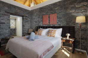 Ein Schlafzimmer mit Steinwand, Holzboden, einem Bett mit weißer Bettwäsche, gemusterten Kissen, einem Nachttisch mit Lampe und einem Beistelltisch.