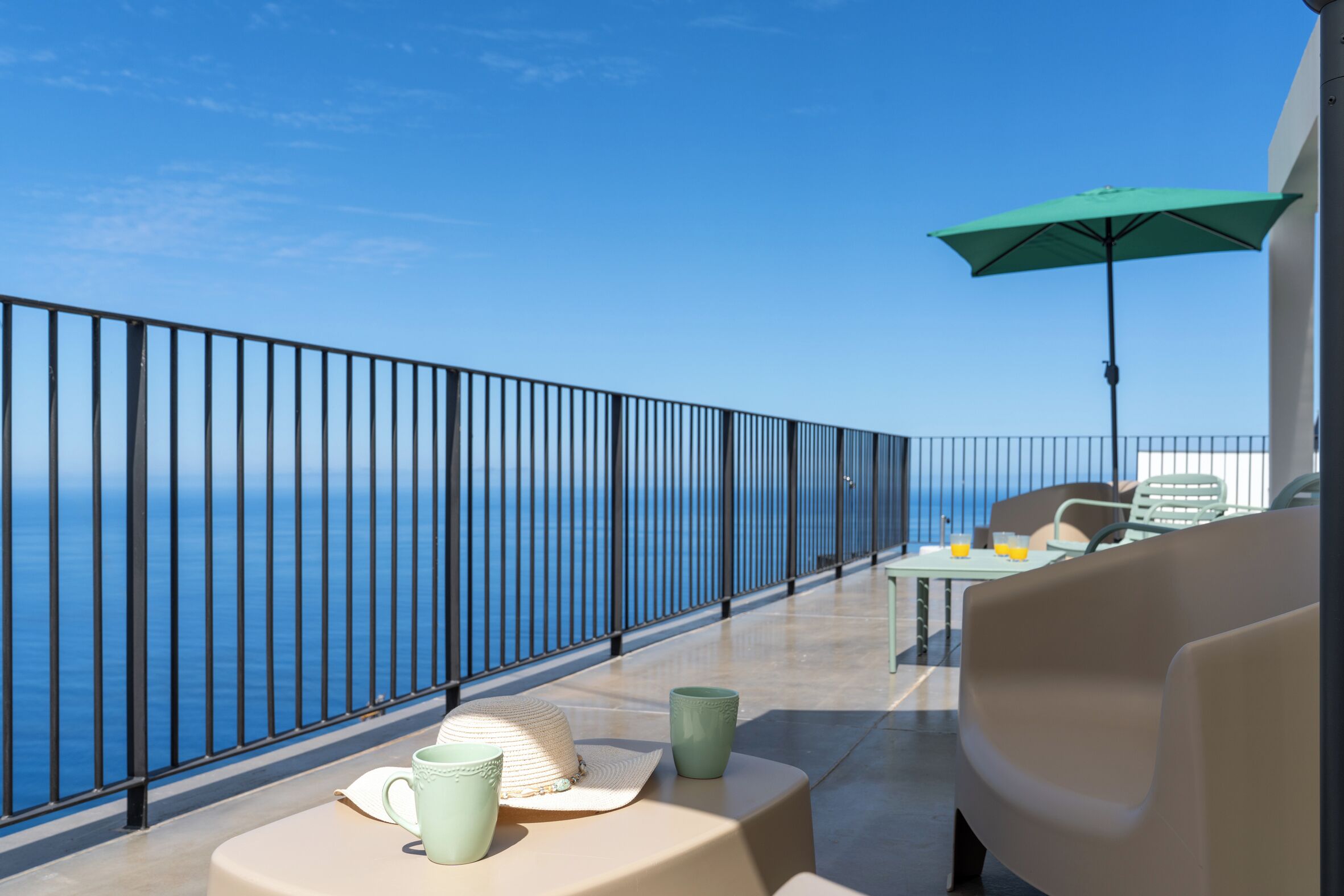 A balcony with a view of the sea, a table with chairs, and a green umbrella.