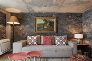 A living room with a stone wall, a sofa with patterned cushions, a painting, and a lamp. - Tranquil with stunning seaview|Ocean Hills Cottage (Faja Da Ovelha)