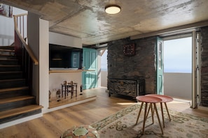 A spacious living area with a wooden staircase, a kitchen with teal cabinets, a stone fireplace, and a large window. - Tranquil with stunning seaview|Ocean Hills Cottage (Faja Da Ovelha)