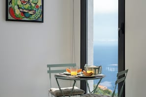 A dining area with a table set for two, a window with a view, and a painting on the wall. - Tranquil with seaview|Ocean Hills Garden Studio (Faja Da Ovelha)