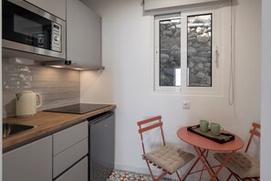 A compact kitchen with a dishwasher, a kettle, and a window with a stone wall view. There is a small dining area with a table and chairs. - Tranquil with seaview|Ocean Hills Balcony Studio (Faja Da Ovelha)