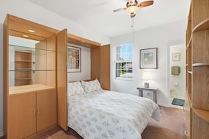 A bedroom with a bed, a wardrobe, a glass-fronted storage unit, a ceiling fan, a window with blinds, and a small bedside table with a lamp.