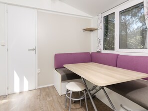 A room with a wooden table, a purple bench, a white stool, a window with a view of trees, and a white door.