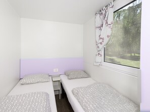 Habitació petita amb dos llits individuals, una finestra amb cortines florals i vista a la verdura a l'exterior.