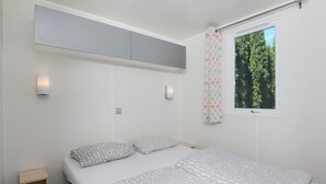 A bedroom with a bed, a small bedside table, a wall-mounted light, a window with a curtain, and a ceiling-mounted storage unit. - Vacation home by Interhome (Osecná)