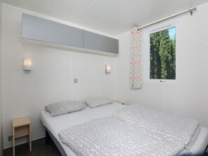 Ein Schlafzimmer mit Bett, einem kleinen Nachttisch, einer Wandleuchte, einem Fenster mit Vorhang und einem Deckenregal.
