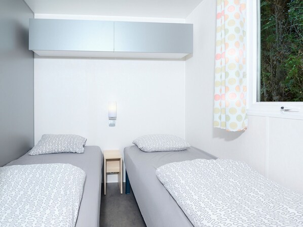 Una habitación con dos camas individuales, una mesita pequeña, una lámpara de pared y un ventanal con cortina.