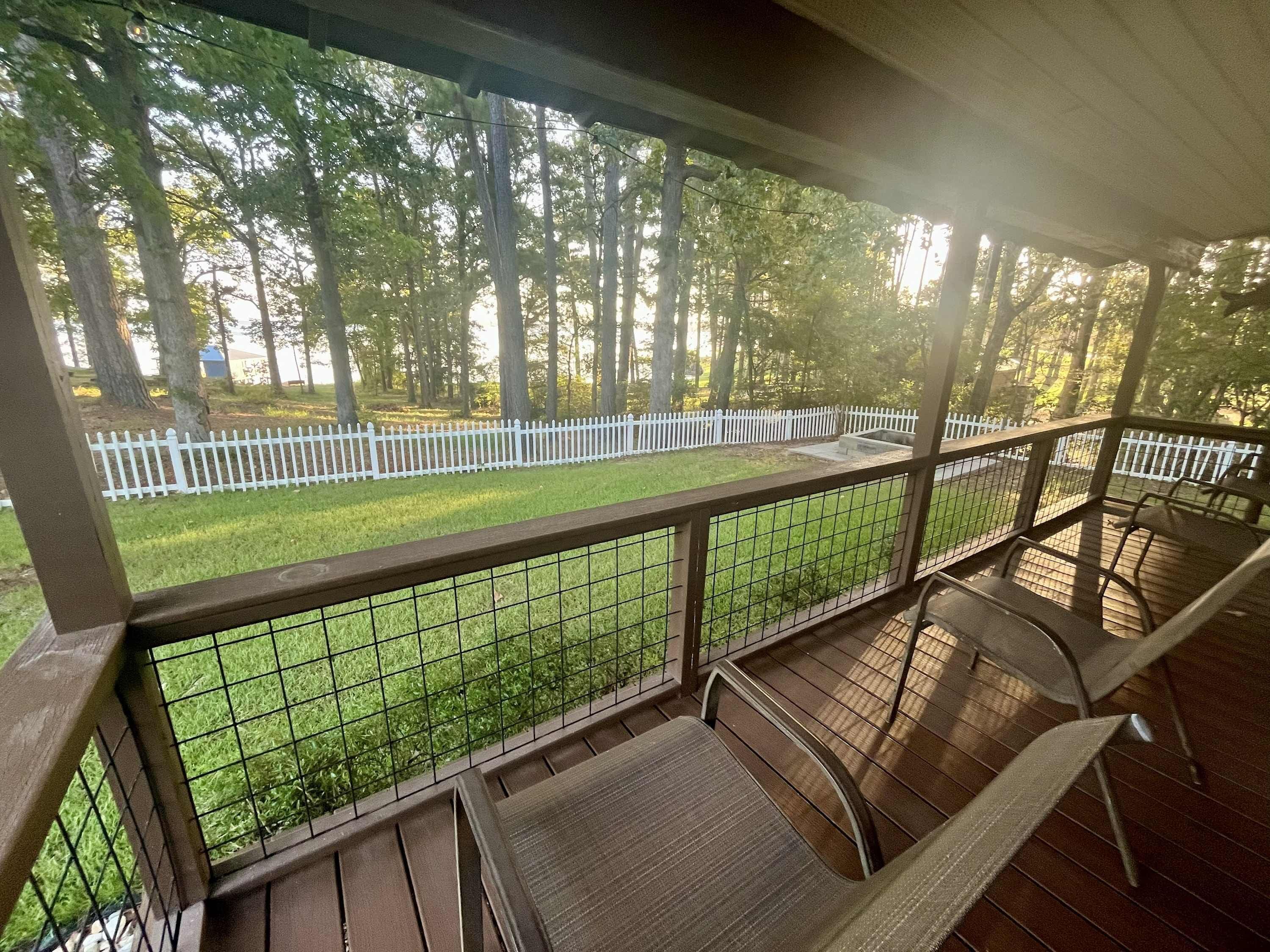 A covered outdoor area with a wooden deck, metal railing, and a view of a white picket fence and trees.