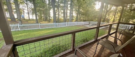 A covered outdoor area with a wooden deck, metal railing, and a view of a white picket fence and trees.