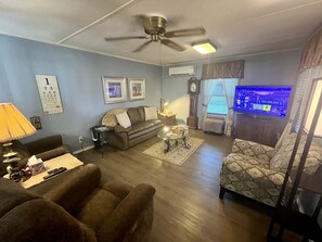 Ein Wohnzimmer mit Deckenventilator, einer Couch, einem gemusterten Sessel, einem Couchtisch und einem Fernseher.
