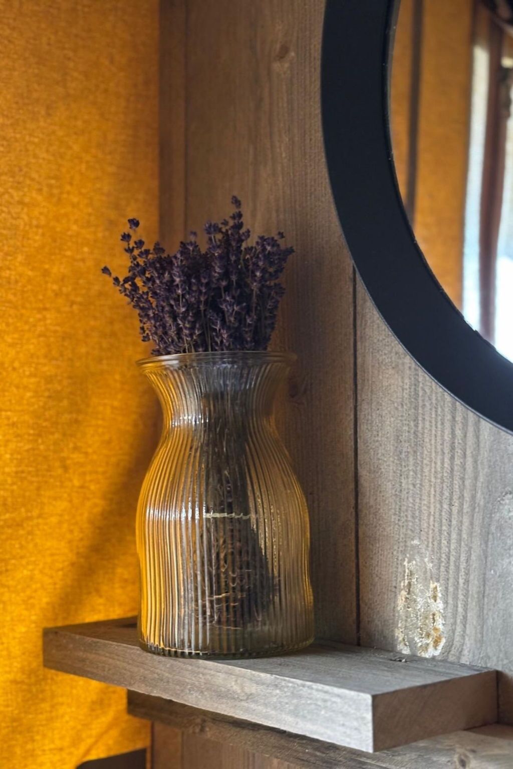 Eine klare Glasvase mit violetten Blumen auf einem hölzernen Regal vor einer strukturierten Wand.