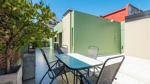 En udendørs terrasse med bord og stole, omgivet af grønt og bygninger.