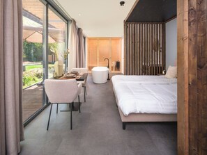 Una camera moderna con un letto grande, una zona salotto con un tavolo e sedie, e vista sull'esterno attraverso ampie finestre.