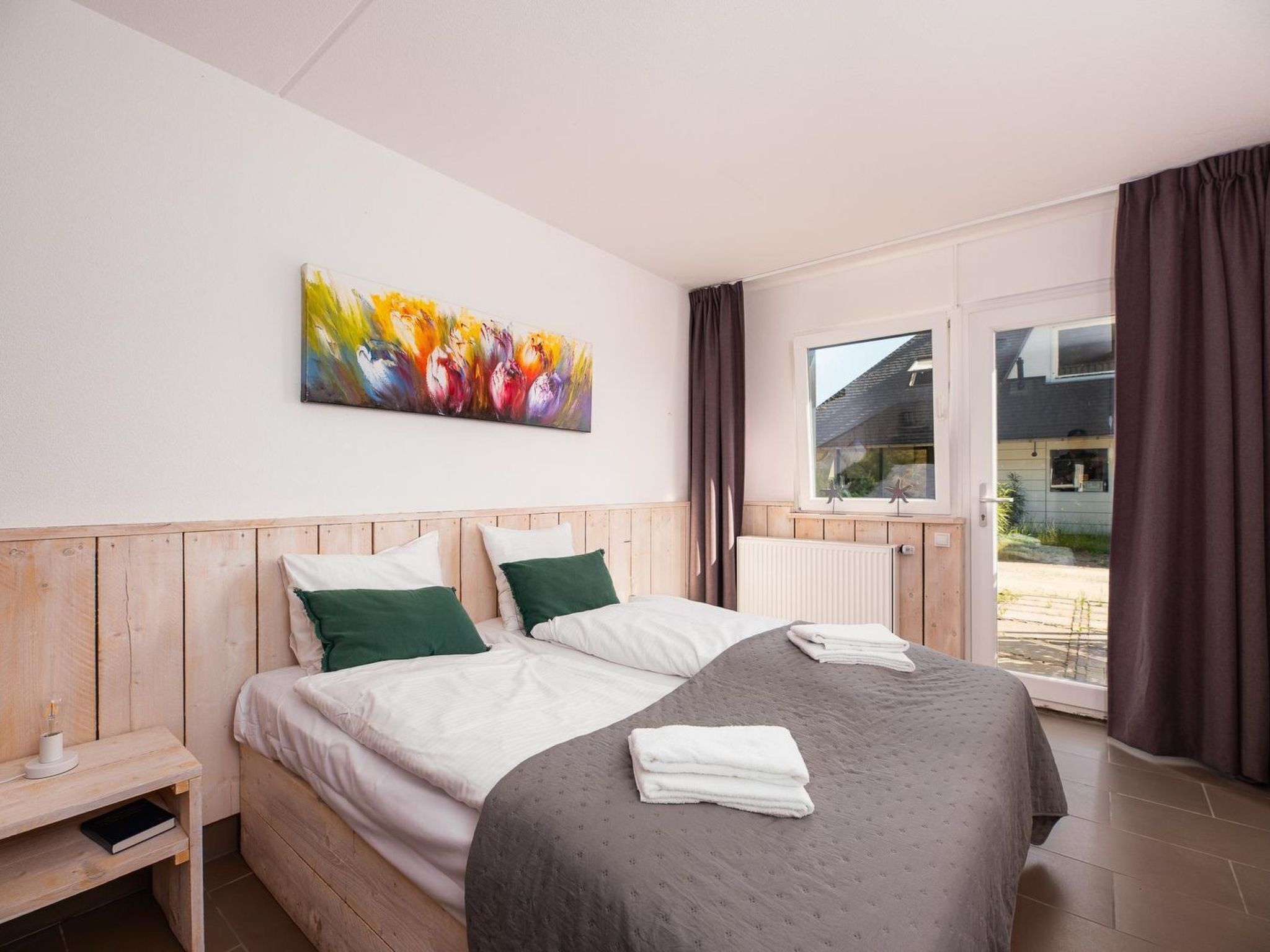 A bedroom with a wooden headboard, a large bed with white and green bedding, a colorful abstract painting on the wall, and a view of the outside through a window with curtains.