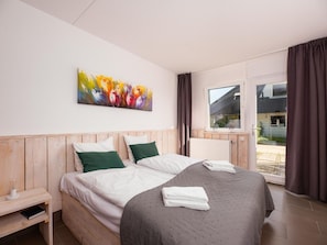 A bedroom with a wooden headboard, a large bed with white and green bedding, a colorful abstract painting on the wall, and a view of the outside through a window with curtains.
