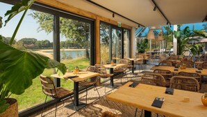 An outdoor dining area with wicker furniture, large windows, and a view of a lake. - Wilsumer Berge Resort - Trekkershut by Interhome (Wilsum)