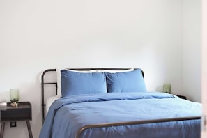 A single bed with a metal frame and a blue bedspread. There is a small bedside table with a green lamp on it.