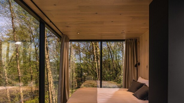 Ein Schlafzimmer mit einem großen Bett, Blick auf Bäume durch raumhohe Fenster und einem modernen Design.