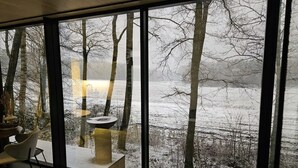 Ein Zimmer mit großem Fenster mit Blick auf eine verschneite Landschaft, Bäume und ein Gewässer.