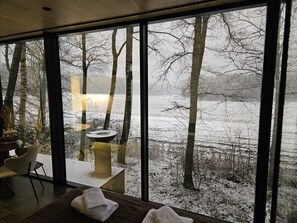Ein Zimmer mit großem Fenster mit Blick auf eine verschneite Landschaft, Bäume und ein Gewässer.
