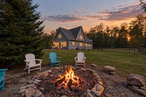 A house with a fire pit in the foreground, surrounded by three Adirondack chairs. - Forest Moon - Peaceful Pet-Friendly Wooded Retreat (Egg Harbor)