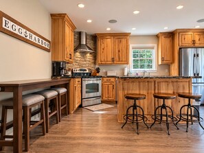 A kitchen with wooden cabinets, a stone backsplash, a stainless steel range hood, a double oven, a refrigerator, and a bar area with stools. - Forest Moon - Peaceful Pet-Friendly Wooded Retreat (Egg Harbor)