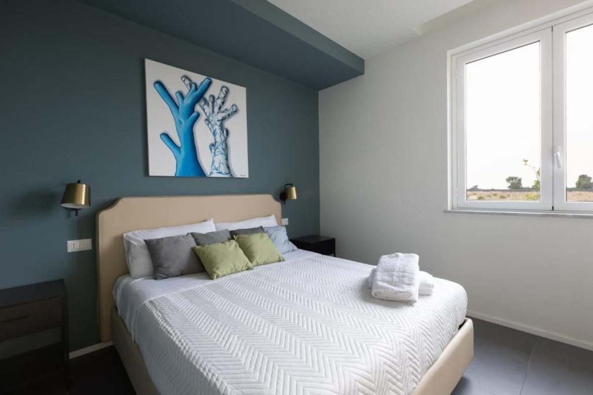 Une chambre à coucher comprenant un lit, une table de chevet, une lampe, une fenêtre et une œuvre d’art murale.