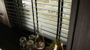 Ein Fenster mit horizontalen Jalousien, ein kleiner Tisch mit Gläsern und eine Pflanze.