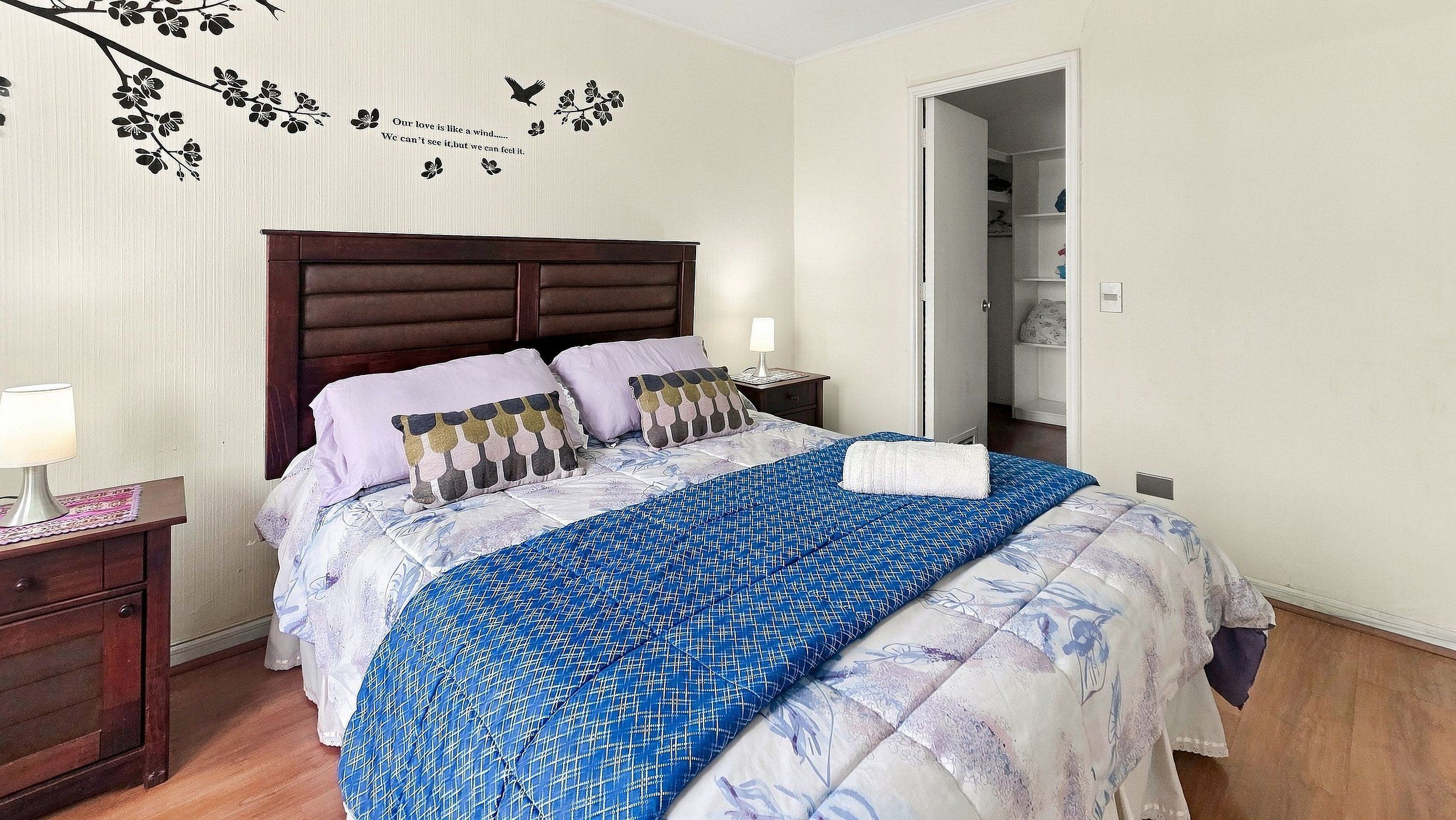 Un dormitorio con cabecera de madera, cama con ropa de cama estampada, mesita de noche con lámpara y una pared con una rama decorativa y texto.