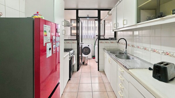 A narrow kitchen with a red refrigerator, a washing machine, and a sink with a faucet.