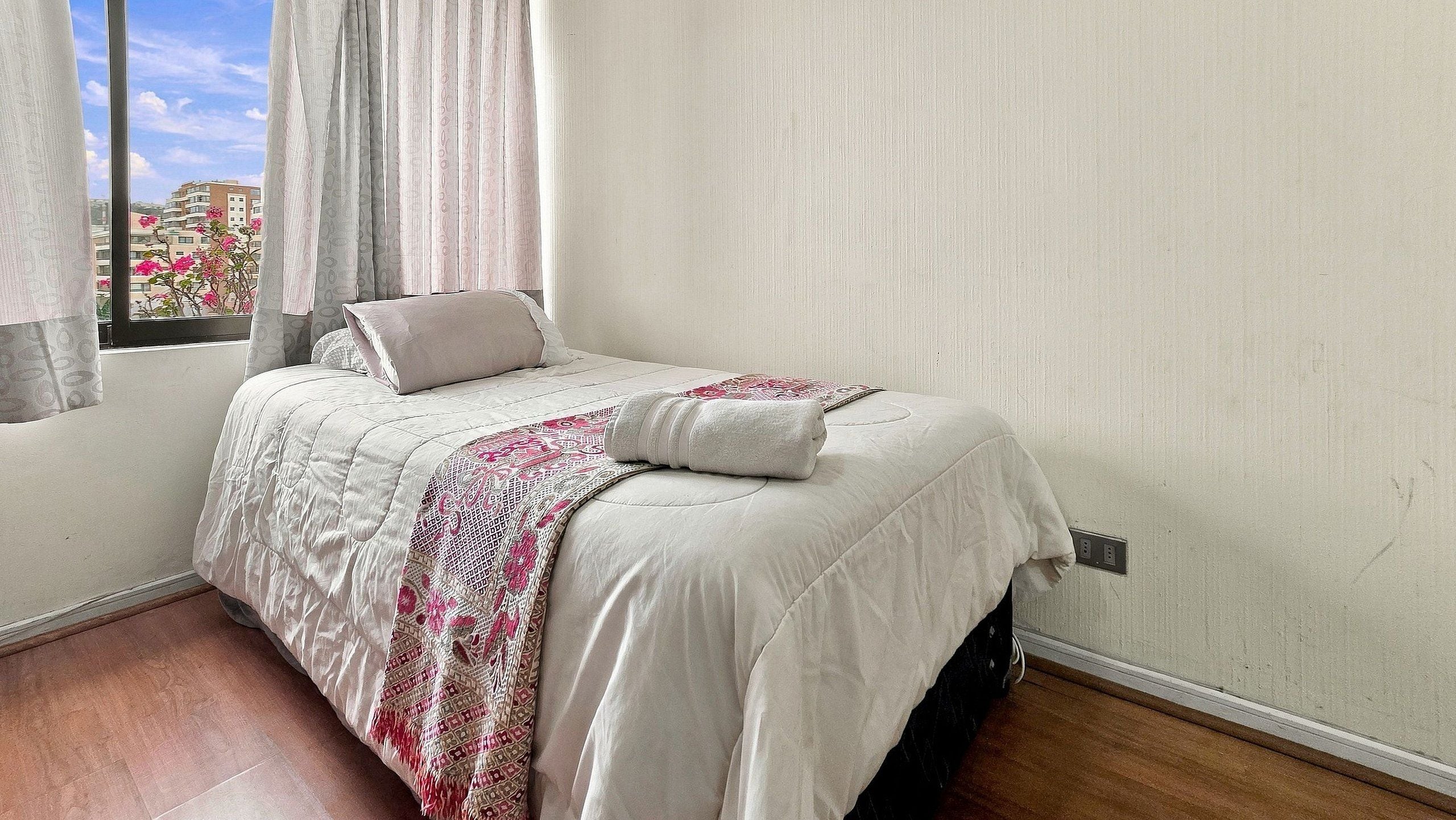 A bedroom with a bed, a window with curtains, and a view of buildings outside.