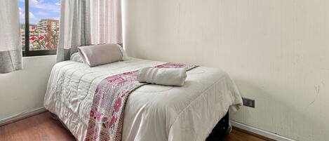 Un dormitorio con cama, ventana con cortinas y vista a edificios.