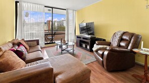 A living room with a sofa, armchair, TV, and a view of buildings through a large window.