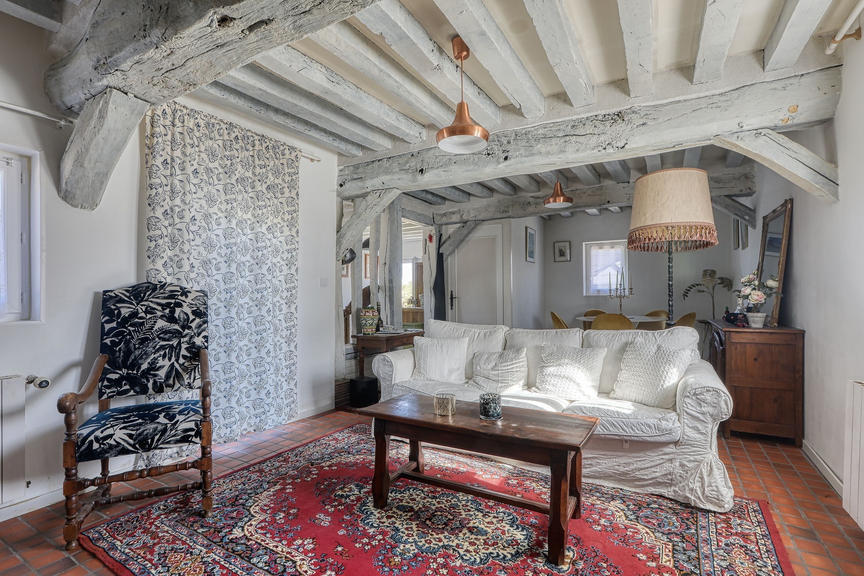 Una habitación con un sofá blanco, un sillón estampado, una mesa de centro de madera y una alfombra roja con diseño.