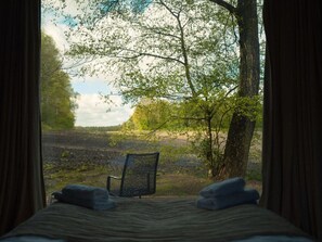 Un lit donnant sur une forêt, avec des rideaux, une chaise et une couverture.