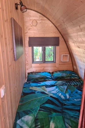 Een kleine, gezellige kamer met een gebogen houten plafond, een raam met donkere gordijnen, en een bed met een sprei met bladprint.