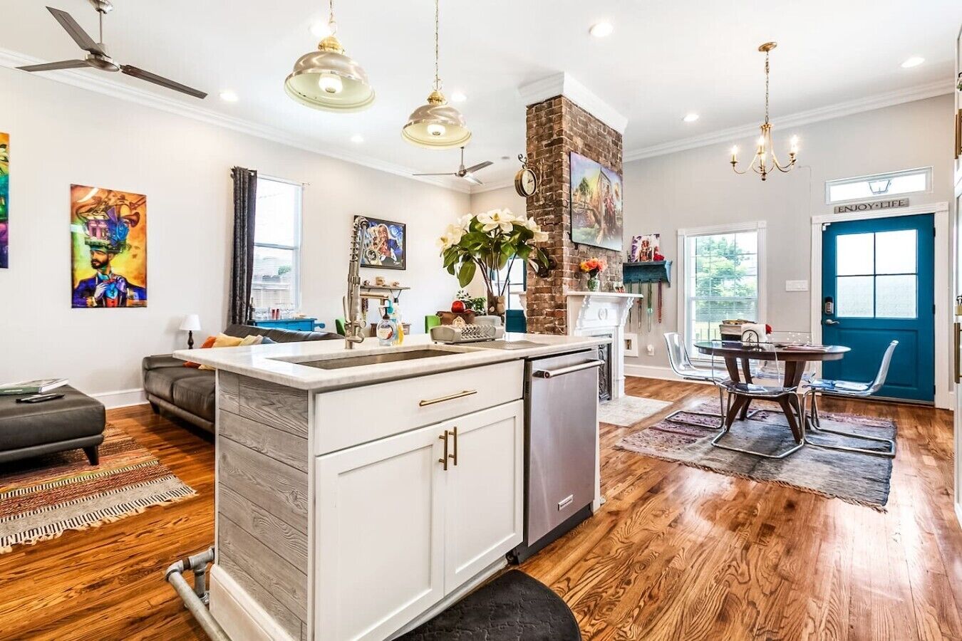 A modern kitchen with a central island, a dining area with a blue door, and a living room with a sofa and artwork.