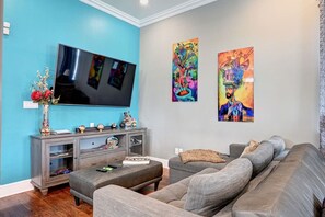 A living room with a flat-screen TV, grey sofa, and two colorful paintings on the wall. - 2BR Home 1 Block to Streetcar, Near French Quarter (New Orleans)