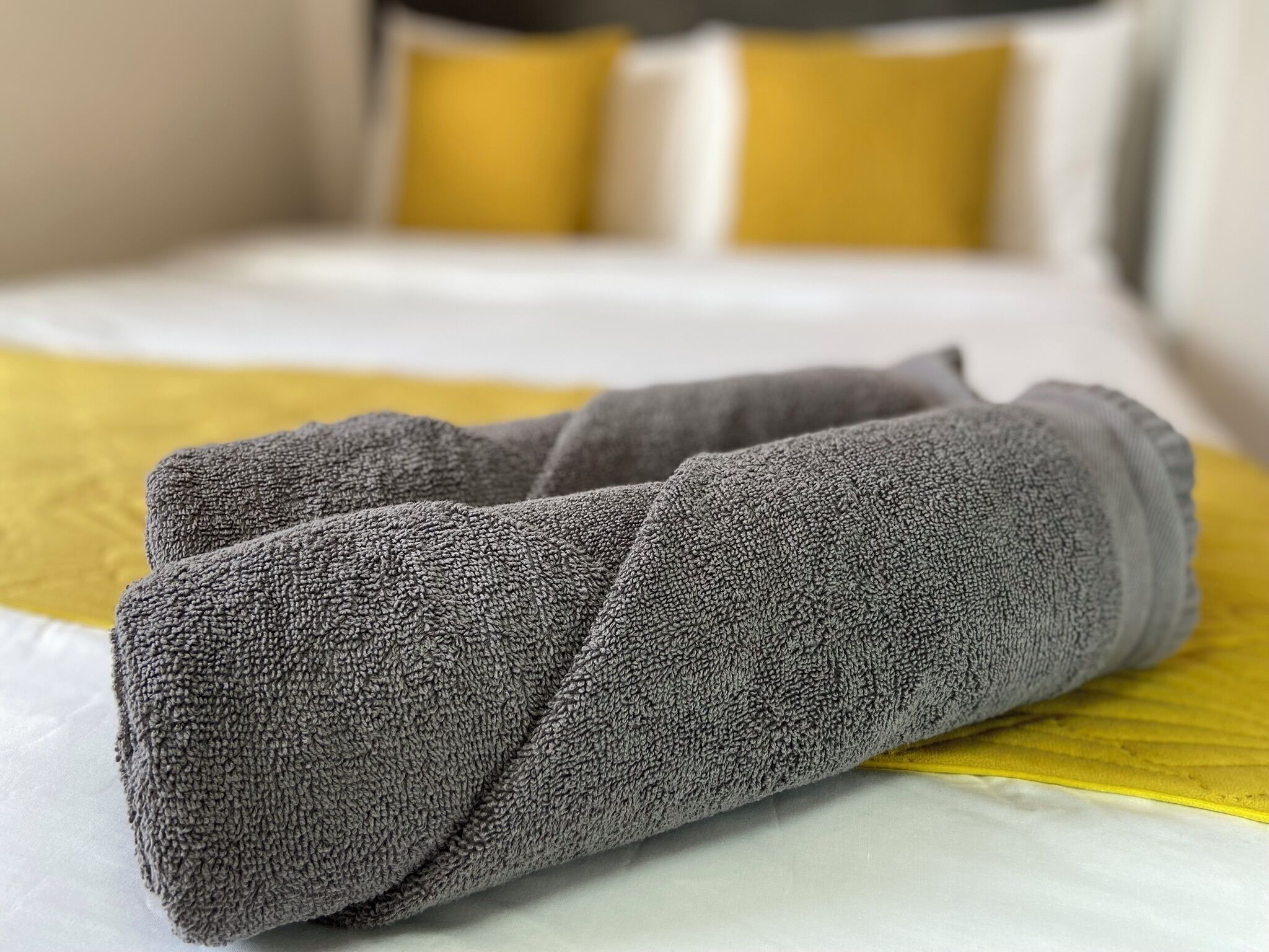 A neatly folded gray towel on a bed with a yellow accent pillow.