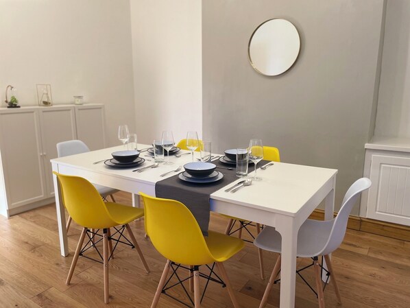 Un coin repas avec une table blanche dressée pour quatre personnes, des chaises jaunes et un miroir rond accroché au mur.