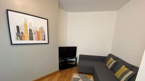 Ein Wohnzimmer mit grauem Sofa, einem schwarzen TV-Schrank, einem Stadtbild im Bilderrahmen und einem gemusterten Teppich.