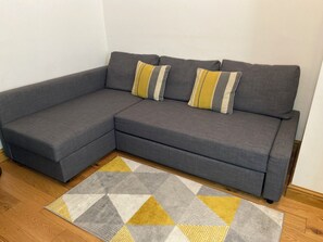 Un canapé d’angle gris avec deux coussins, un tapis à motifs géométriques et un plancher en bois.