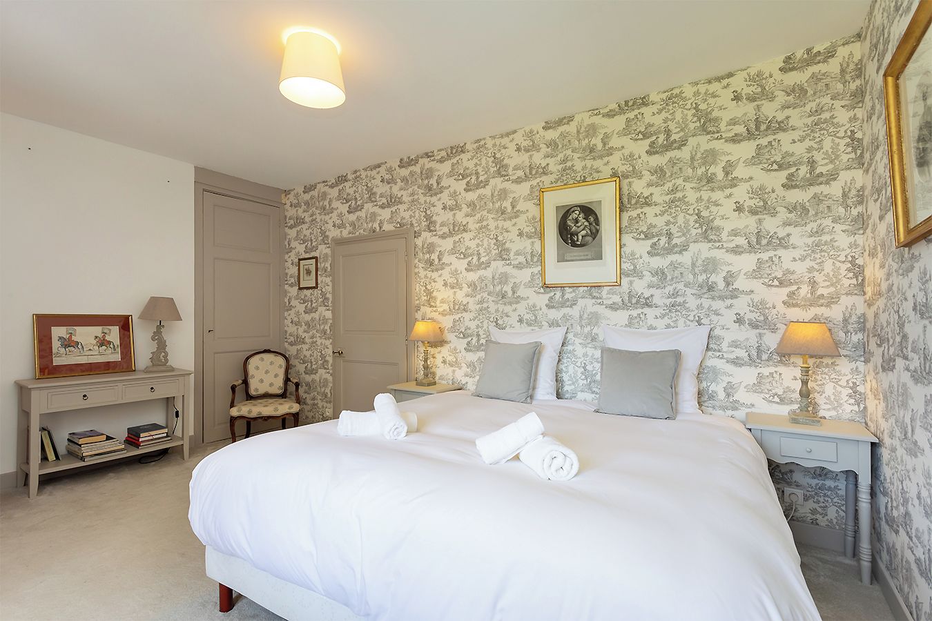 Un lit bien fait, avec des oreillers et une serviette enroulée, une chaise, une table de chevet avec une lampe et un tableau encadré au mur.