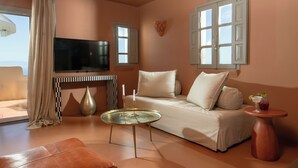A living room with a sofa, a coffee table, a TV, and a window with shutters. - Terra Verde Oia Suites - Terracote Grand Suite Sea View with Outdoor Jetted Tub (Oia)