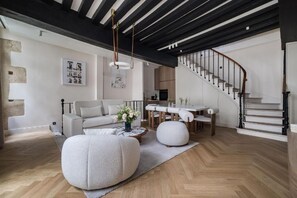 A modern living room with a sofa, ottoman, and coffee table. A staircase with a railing is visible in the background. - Parisian Elegance: Stunning 3BR Retreat in the Heart of Le Marais (Paris)