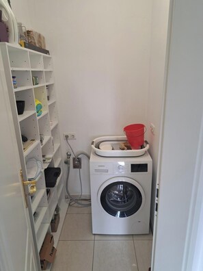 A laundry room with a white washing machine, a shelf with various items, and a red bucket. - Weener Penthouse Right on the Harbor (Weener)