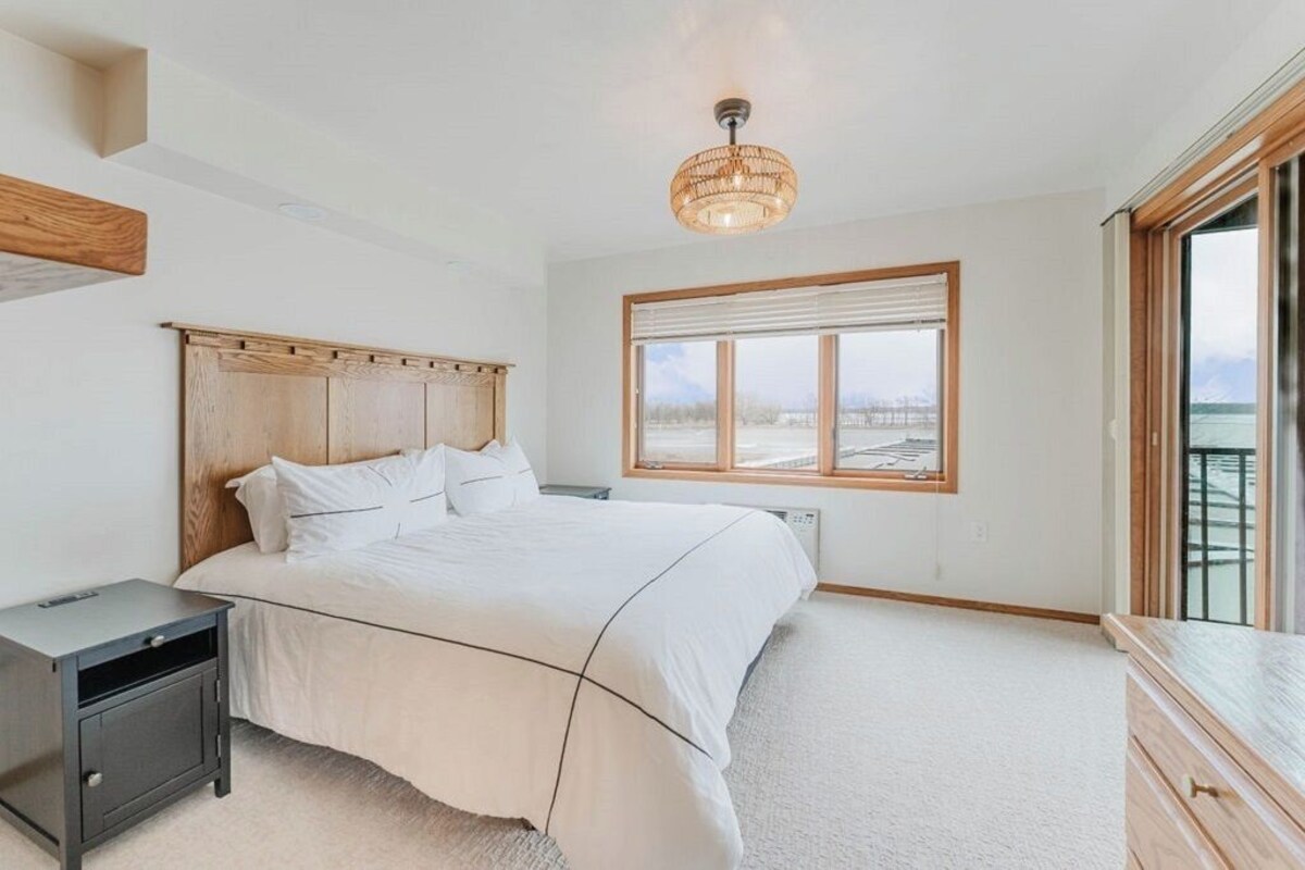 A bedroom with a large bed, a bedside table, a nightstand, a window with blinds, and a view of the outdoors.